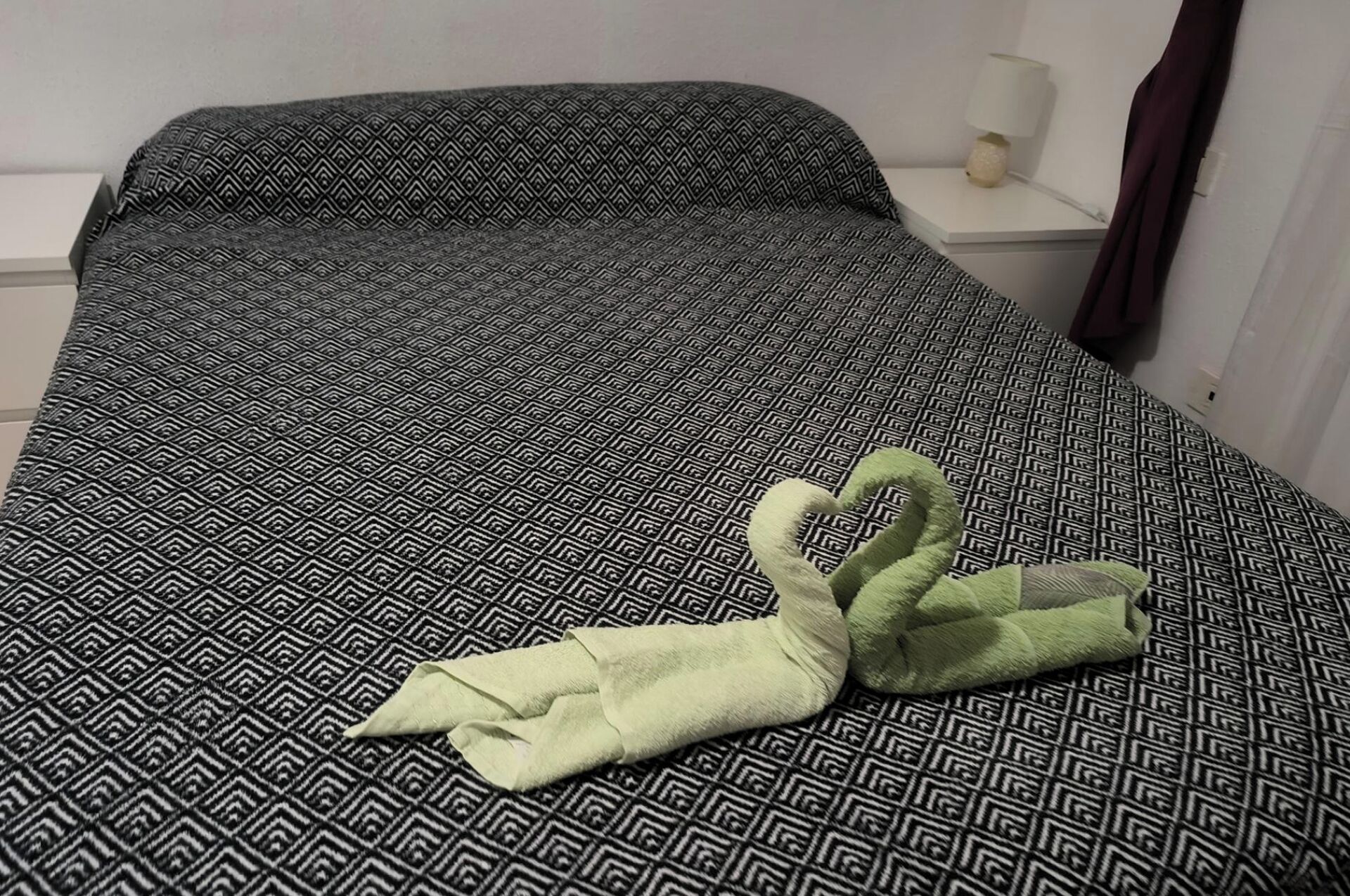 A bed with a geometric patterned bedspread, a white bedside table with a lamp, and a green towel folded on the bed.