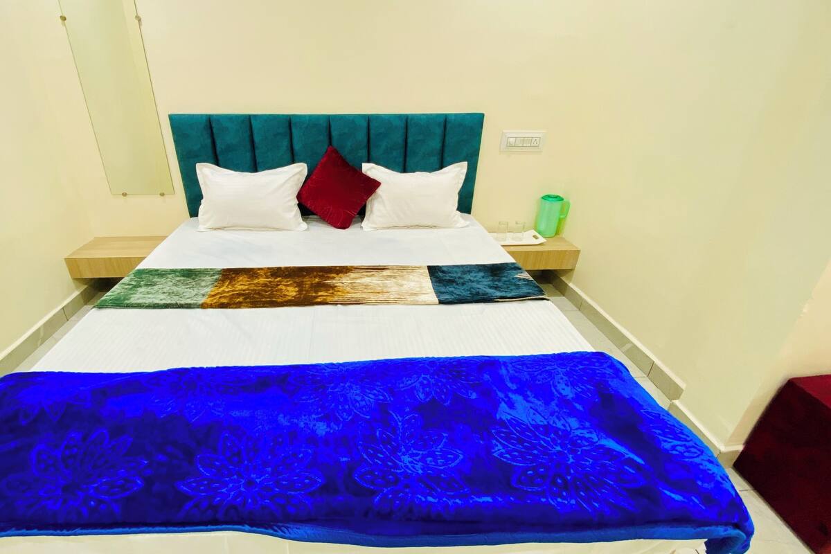 A neatly made bed with a blue floral bedspread, two white pillows, and a red accent pillow. There is a green water bottle on a nightstand beside the bed.