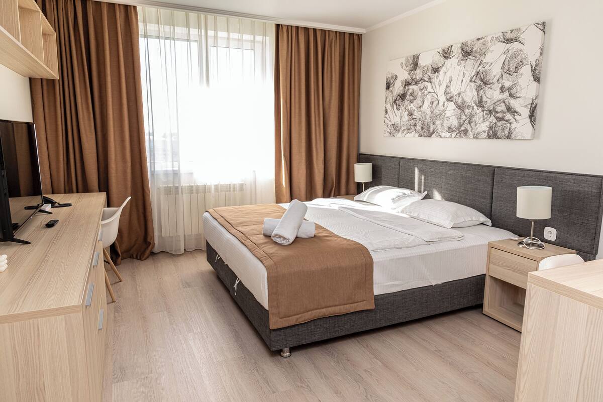 A hotel room with a bed, bedside tables, a desk, and a large window with curtains.