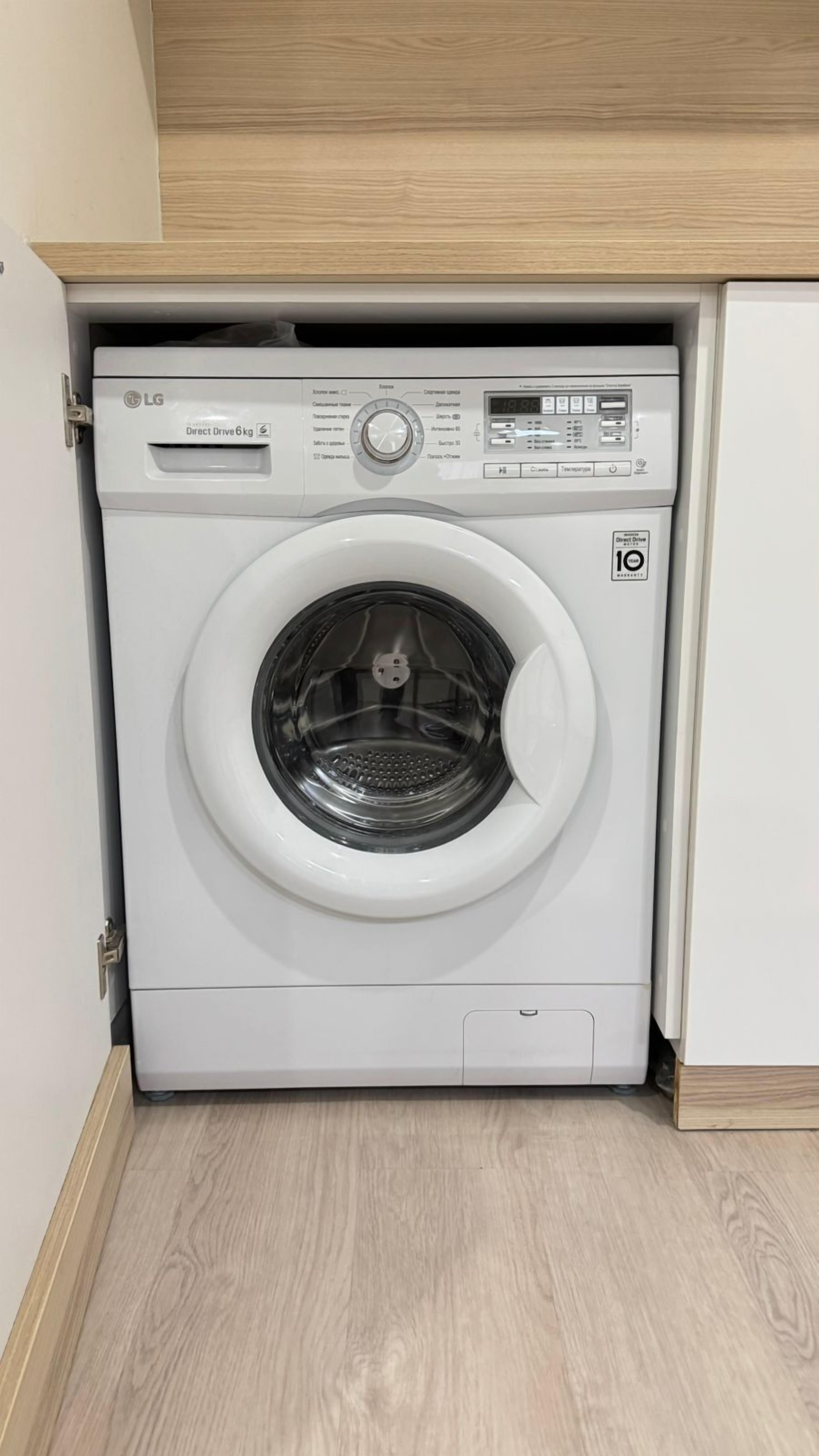 A compact, white LG Direct Drive 6kg front-loading washing machine with a control panel, set in a small laundry room with light wood flooring and a wooden cabinet above.