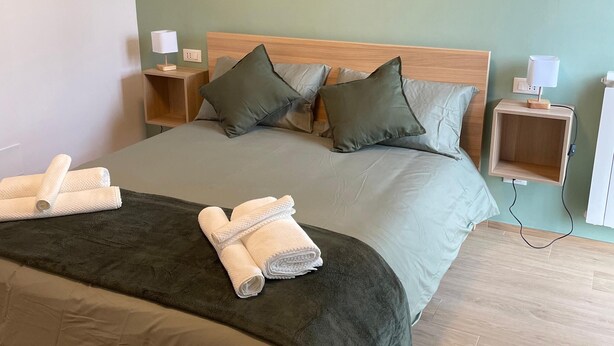 Een net opgemaakt bed met een donkergroene dekbedovertrek en twee kussens, een houten hoofdbord en een aan de muur hangend abstract schilderij.