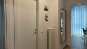 Een gang met een witte deur, een radiator en een spiegel die een kamer met een stoel en een tafel weerkaatst.