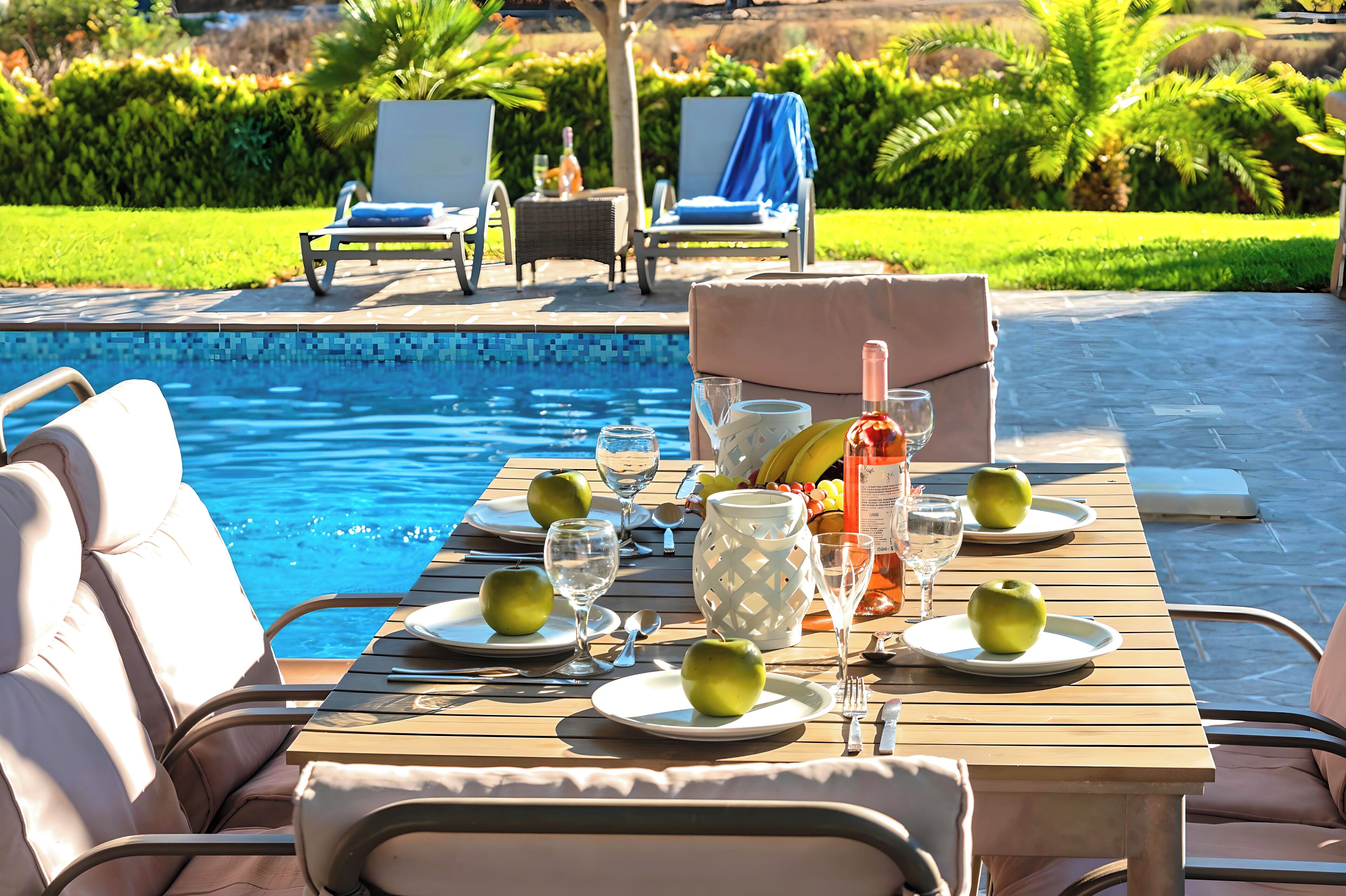 A poolside dining area with a table set for two, surrounded by lounge chairs, a pool, and a garden view.
