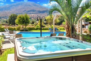 A hot tub with a pool and lounge chairs, surrounded by greenery and a mountain view.