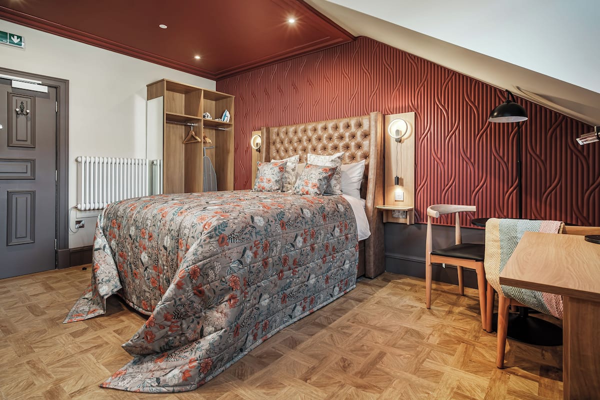 Un dormitorio con cabecera capitonada, un armario de madera, un radiador, una mesa de comedor de madera con sillas y una colcha estampada.
