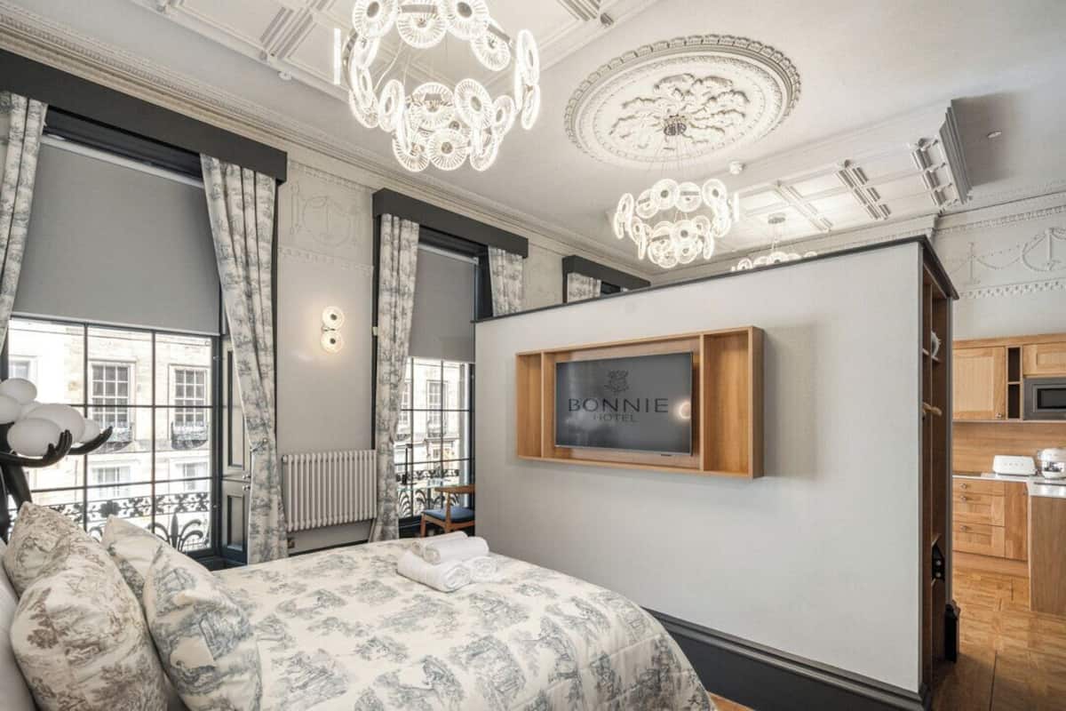 Habitación de hotel con cama, televisión montada y vista de la ciudad a través de grandes ventanales.