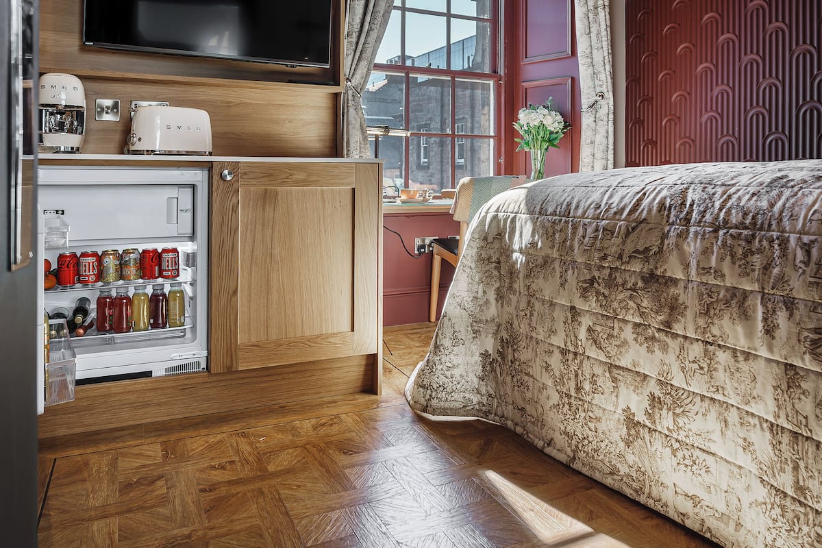 Habitación de hotel compacta con suelo de madera, cama con cubrecama floral, refrigerador empotrado con bebidas, una mesita con una silla y ventana con vistas a edificios.