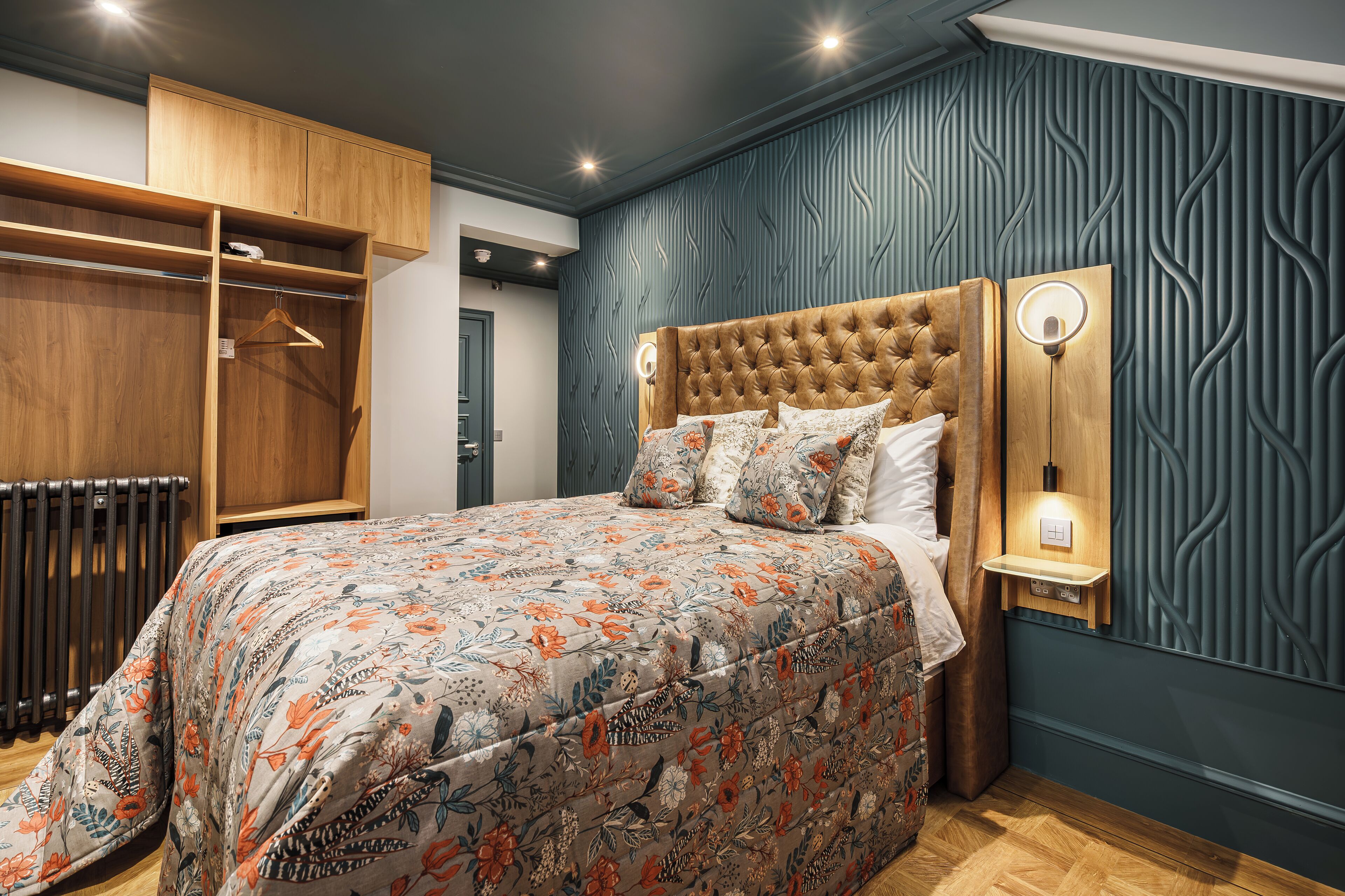 A bedroom with a tufted headboard, floral bedspread, wooden wardrobe, and a wall-mounted nightstand with a light.