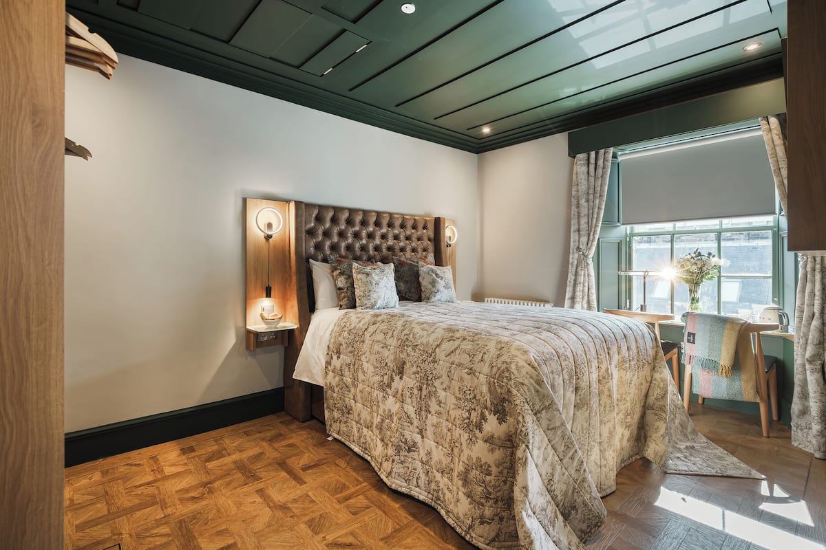 Un dormitorio con cabecera capitonada, una cama grande con colcha estampada, suelo de madera, un escritorio con silla y una ventana con cortinas.