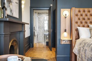 A hotel room with a wooden floor, a fireplace, a bed with a tufted headboard, a bedside table with a lamp, and a view of the bathroom.