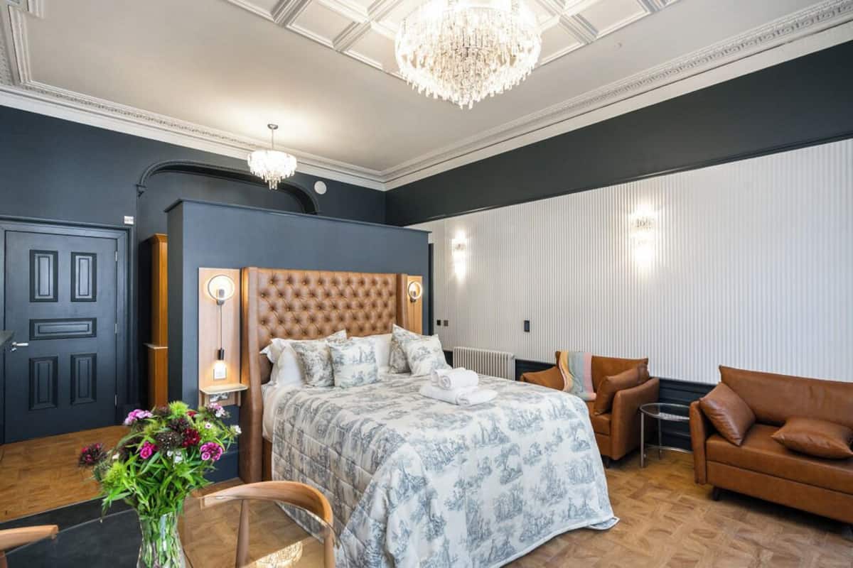 Un dormitorio con cabecera capitonada, una cama grande, una zona de estar con sofá de cuero marrón y un candelabro.