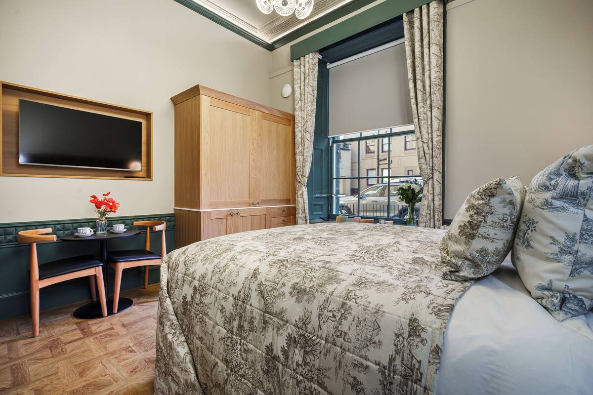 Un dormitorio con cama, armario de madera, escritorio con sillas, ventana con cortinas y vista a la calle.