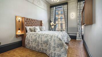 Un dormitorio con cabecera capitonada, colcha estampada, mesita de noche con lámpara, radiador y ventana con cortinas.
