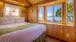 A wooden cabin bedroom with a large bed, a window with a view of the sea, and a framed picture on the wall. - 2BR Deluxe Beachfront Cabin on Spectacular West Beach! (Eastsound)