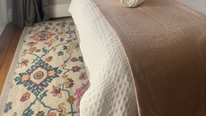 A neatly made bed with a patterned rug, a window with curtains, and a bedside lamp.