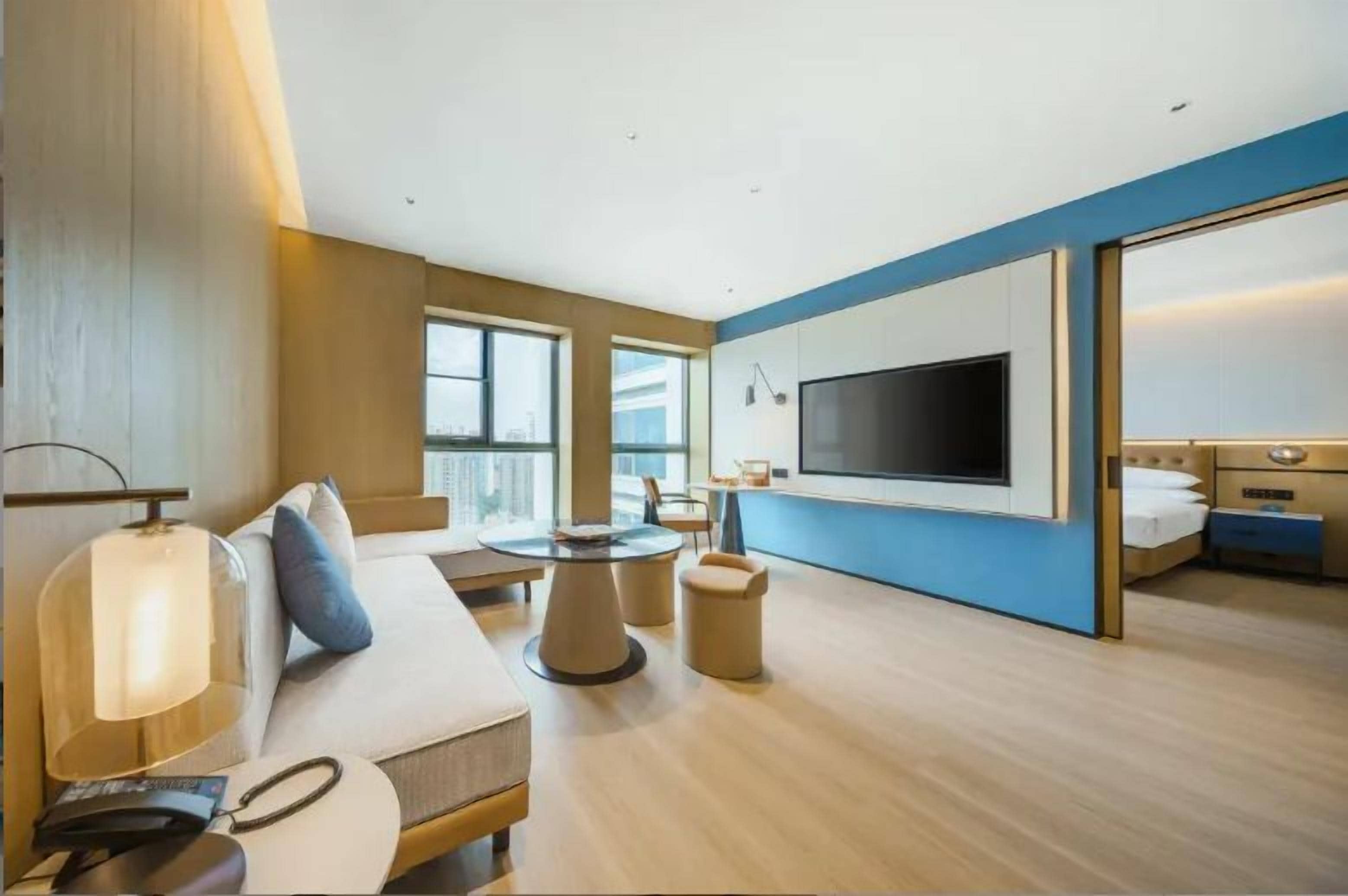 Ein modernes Hotelzimmer mit einem großen Bett, einem flachbildfernseher an der Wand, einer Sitzecke mit einem runden Tisch und Blick auf die Stadt durch große Fenster.