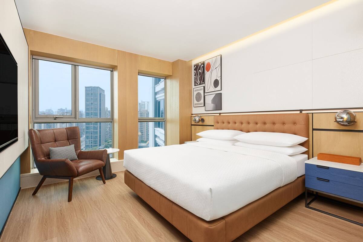 Un hotel moderno con una cama grande, un sillón de cuero, una mesita de noche y vistas de la ciudad a través de amplios ventanales.