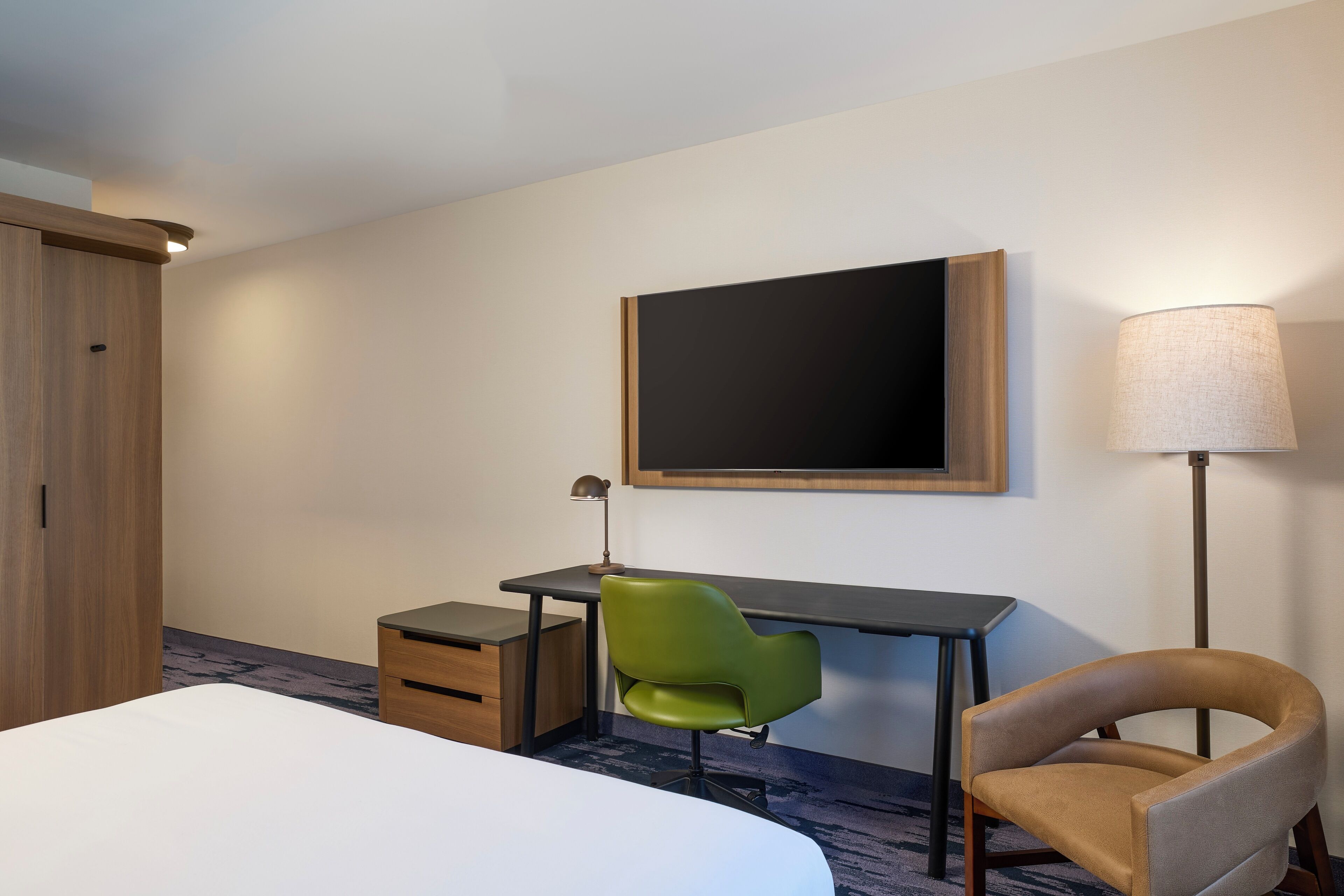A hotel room with a bed, a desk, a green chair, a lamp, and a wardrobe.