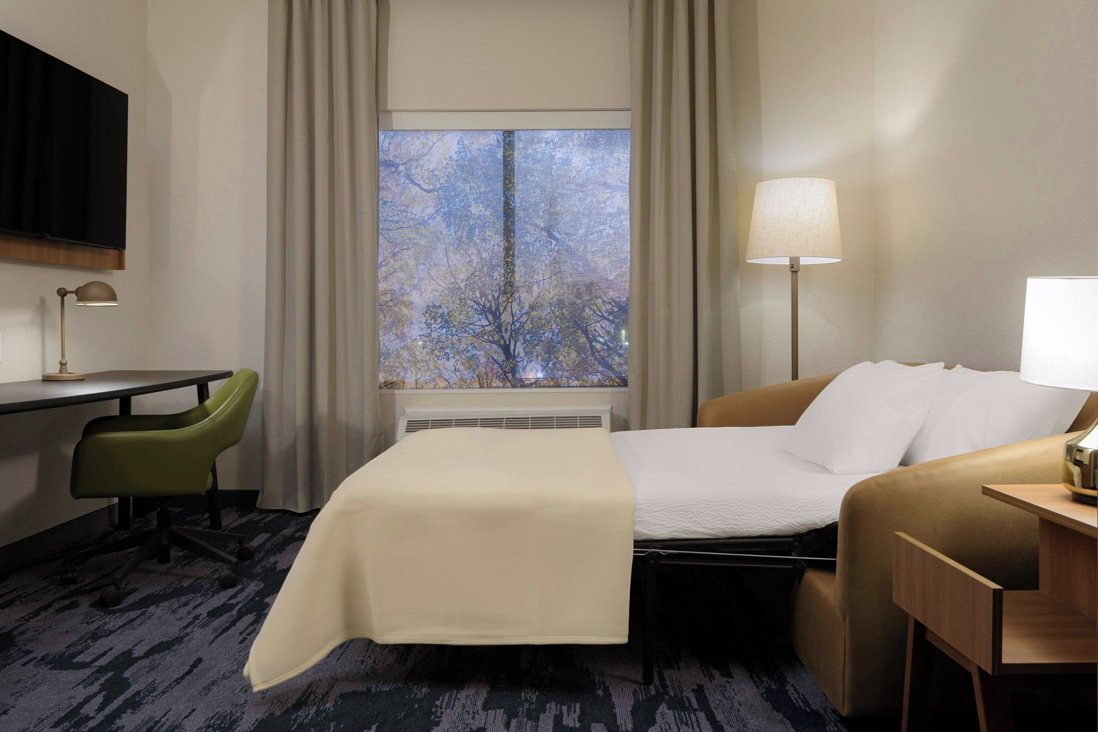 A hotel room with a bed, a desk, a chair, a lamp, and a window with a view of trees.