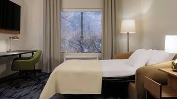 A hotel room with a bed, a desk, a chair, a lamp, and a window with a view of trees.