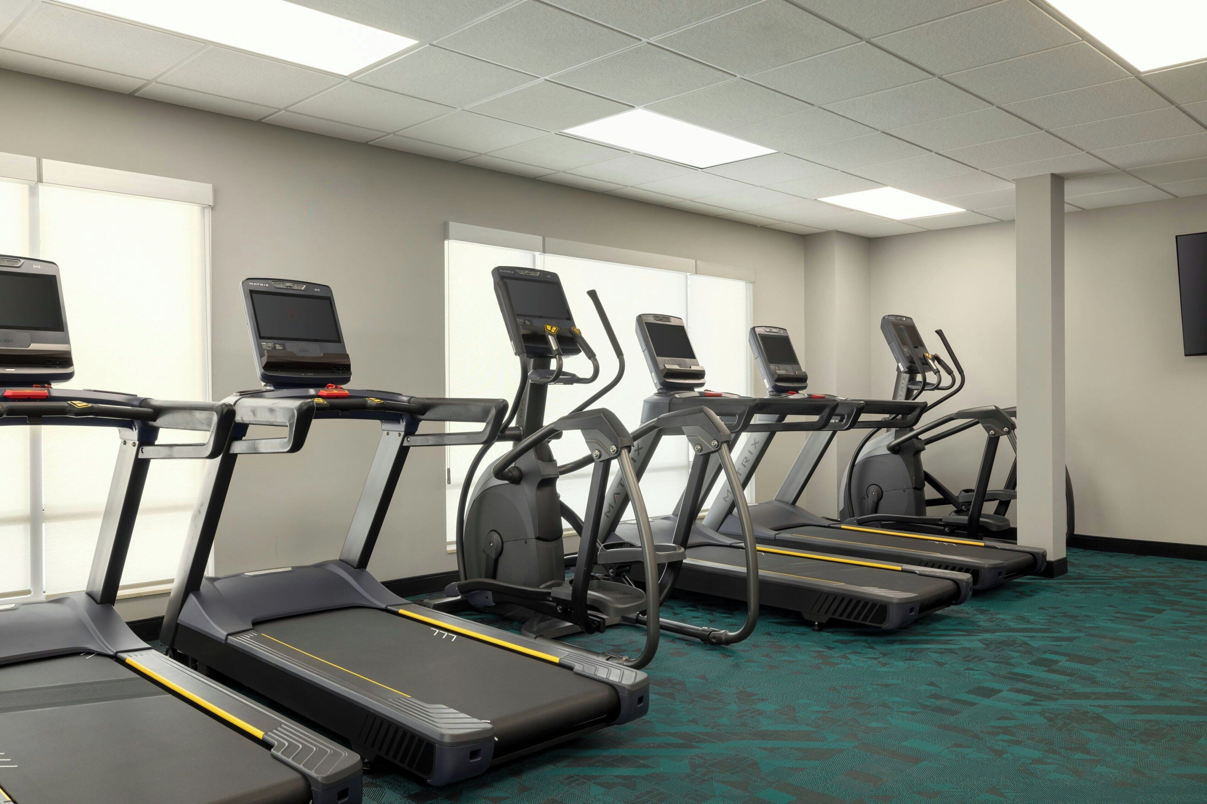 A fitness room with treadmills and elliptical machines, equipped with digital screens.