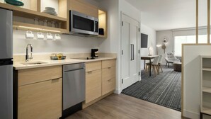 Een moderne keuken met houten kastjes, een RVS vaatwasser en een magnetron. Er is een eethoek met een tafel en stoelen, en een zwart tapijt op de vloer.