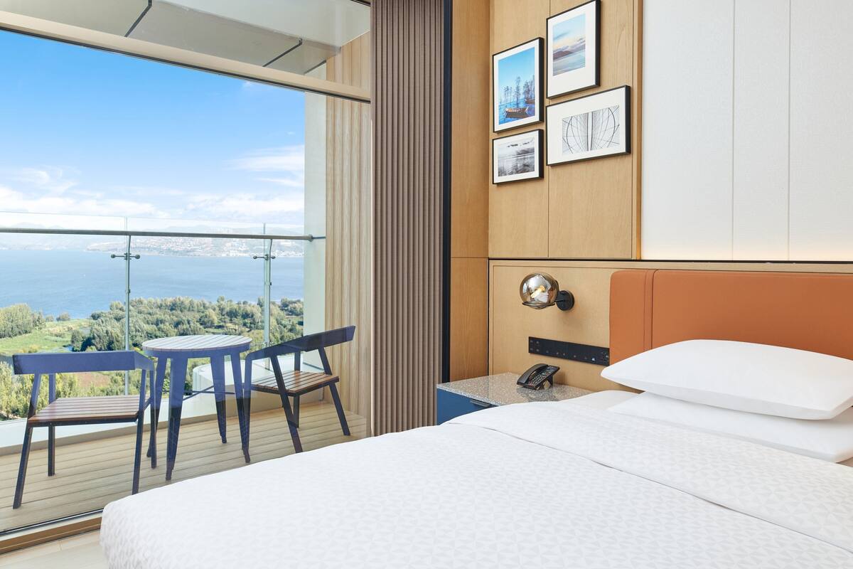 A hotel room with a bed, a small table, two chairs, a phone, and a view of the sea and landscape.