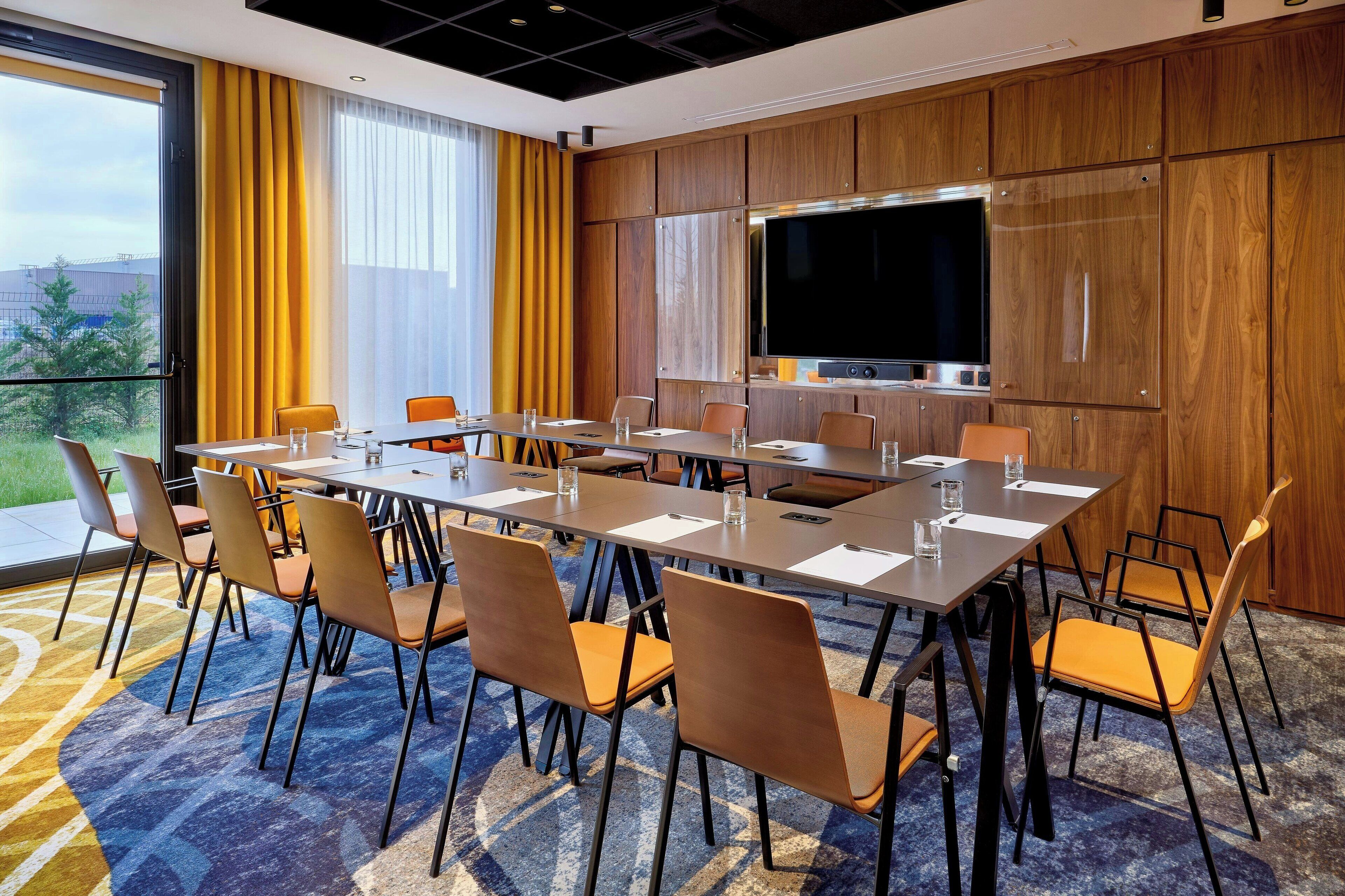 Un salón de conferencias con una mesa grande, sillas, una televisión y vistas al exterior con vegetación.