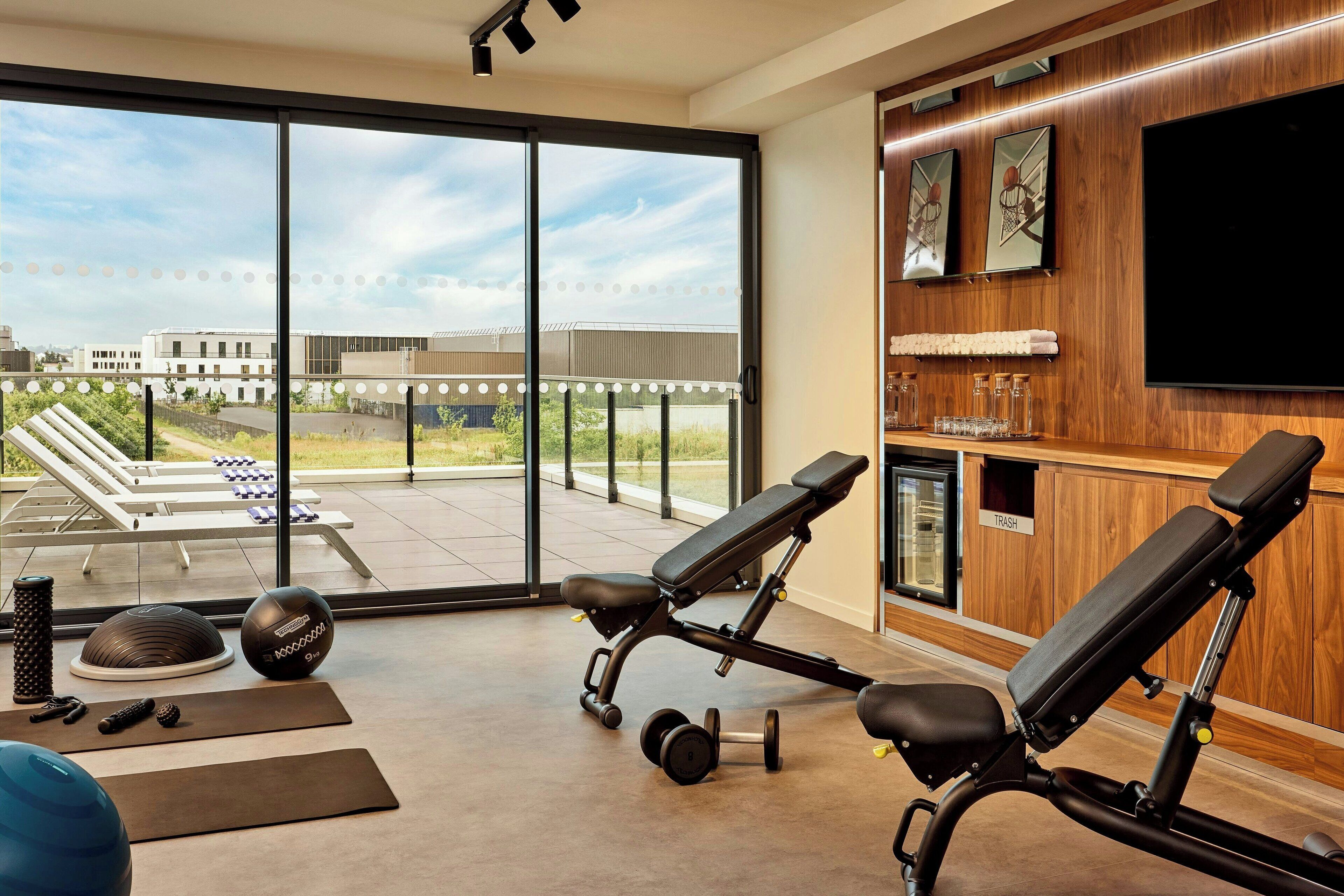 Un gimnasio moderno con equipamiento de ejercicio, ventanas amplias y vistas a una terraza.