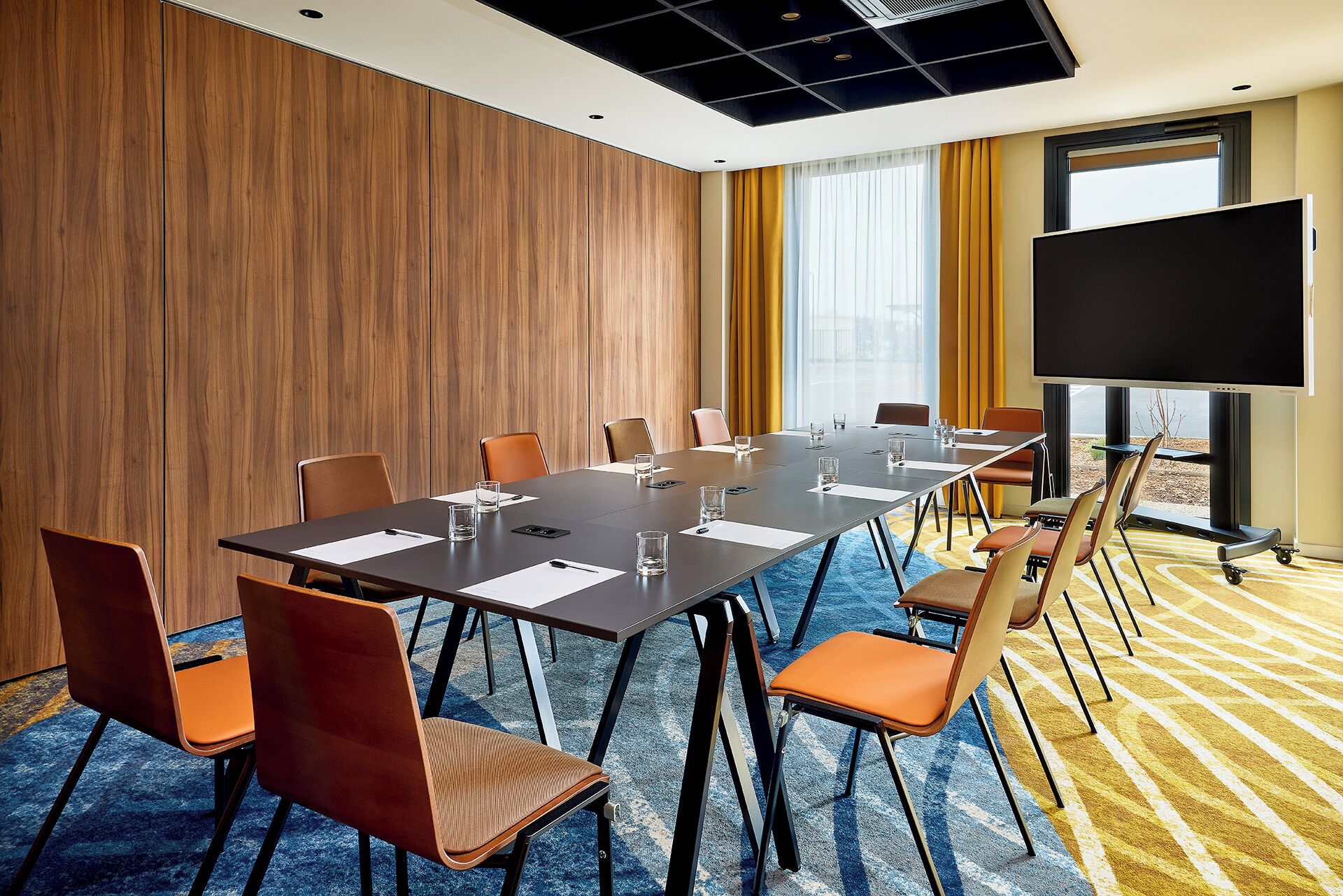 A conference room with a long table, multiple chairs, a large screen, and a view through the window.
