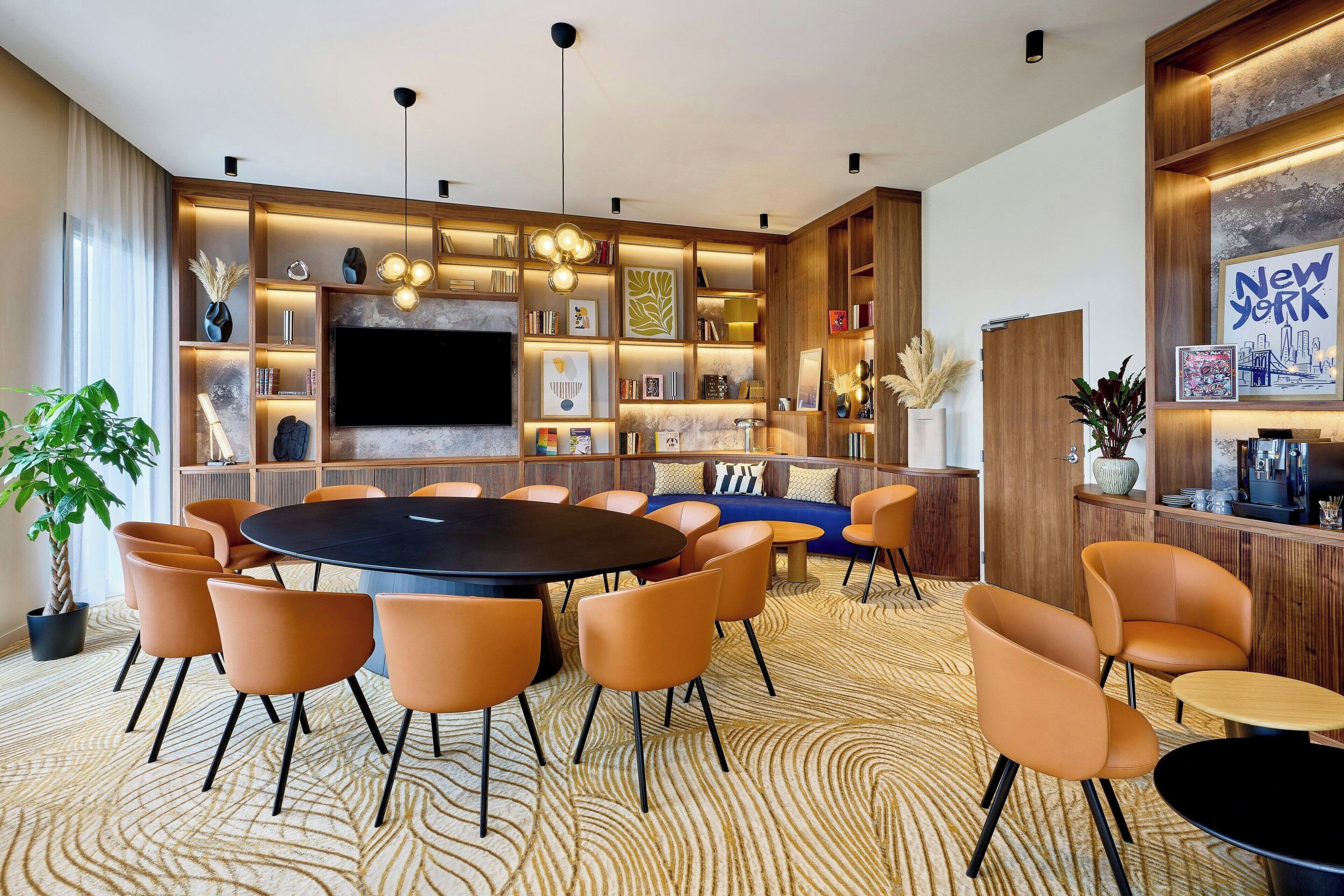 A modern conference room with a round table, orange chairs, and a large TV screen.