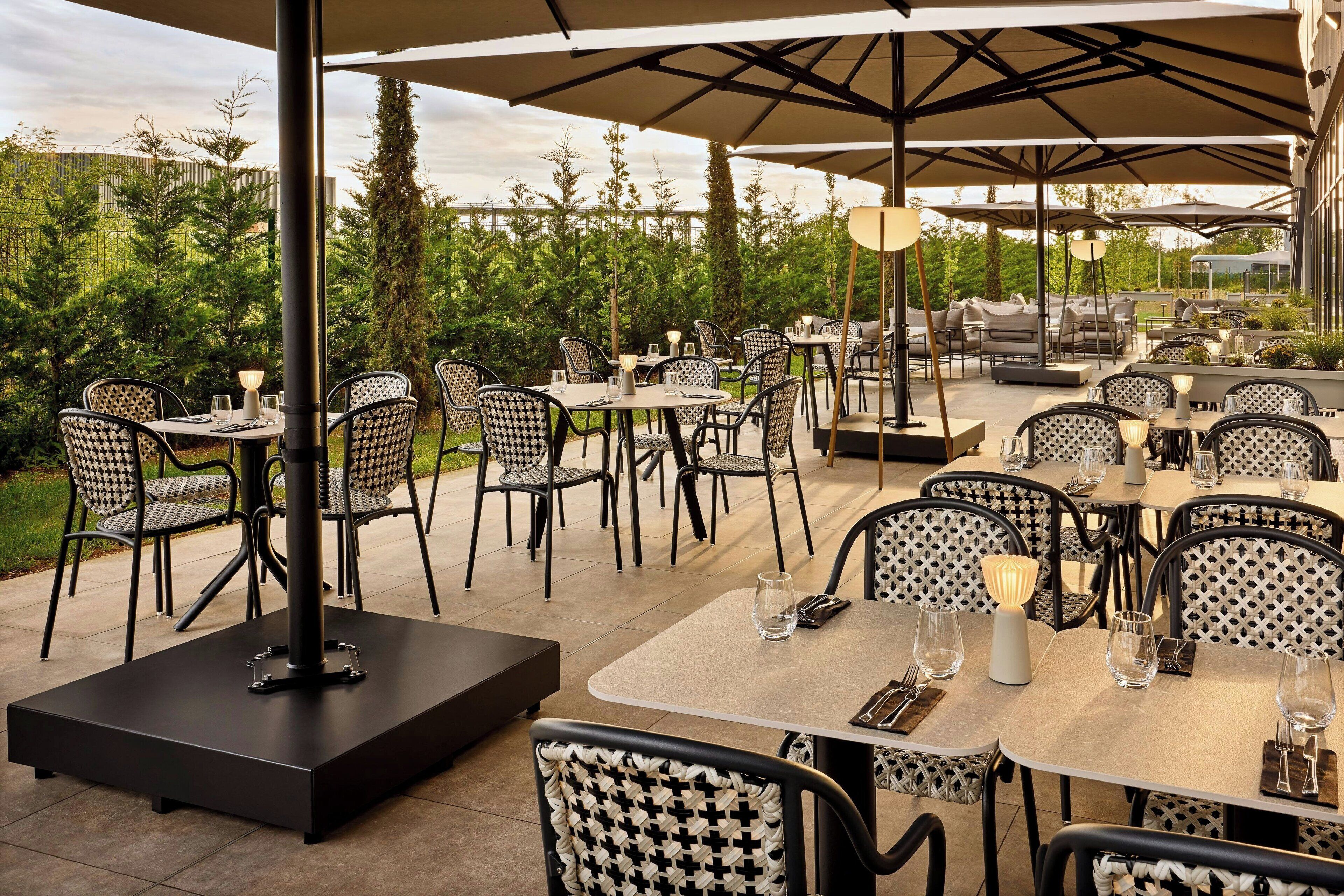 An outdoor dining area with multiple tables and chairs, surrounded by greenery and umbrellas.