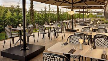 An outdoor dining area with multiple tables and chairs, surrounded by greenery and umbrellas.
