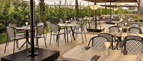 An outdoor dining area with multiple tables and chairs, surrounded by greenery and umbrellas.