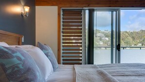 A bedroom with a bed, pillows, a nightstand, a window with blinds, and a view of a body of water and buildings.