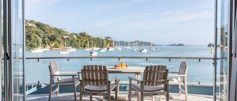 Une terrasse en bois dotée d’une balustrade en verre, qui offre une vue sur une marina avec des bateaux et un paysage vallonné en arrière-plan.