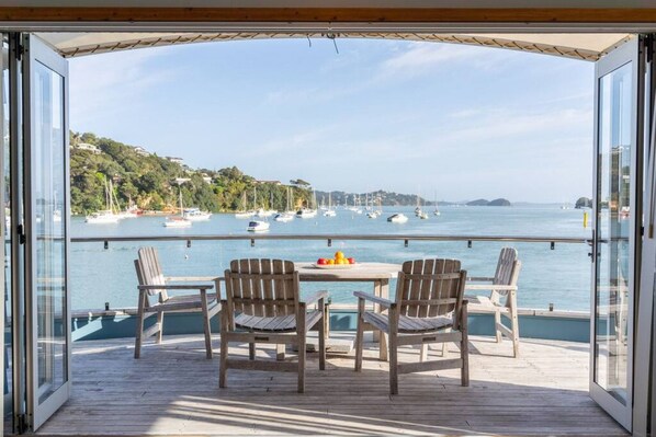 A wooden deck with a glass balustrade overlooking a marina with boats and a hilly landscape in the background.
