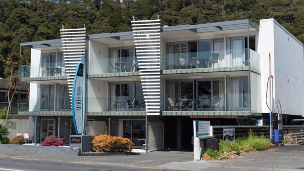 Un edificio moderno de dos pisos con balcones, un letrero visible que dice 'Sea Spray', y vistas de una ladera boscosa.
