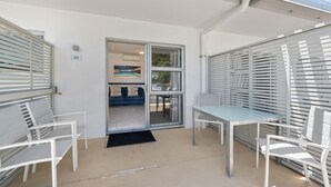 Un patio iluminado por el sol con un ventanal que da a una sala de estar, muebles blancos y vistas al exterior.