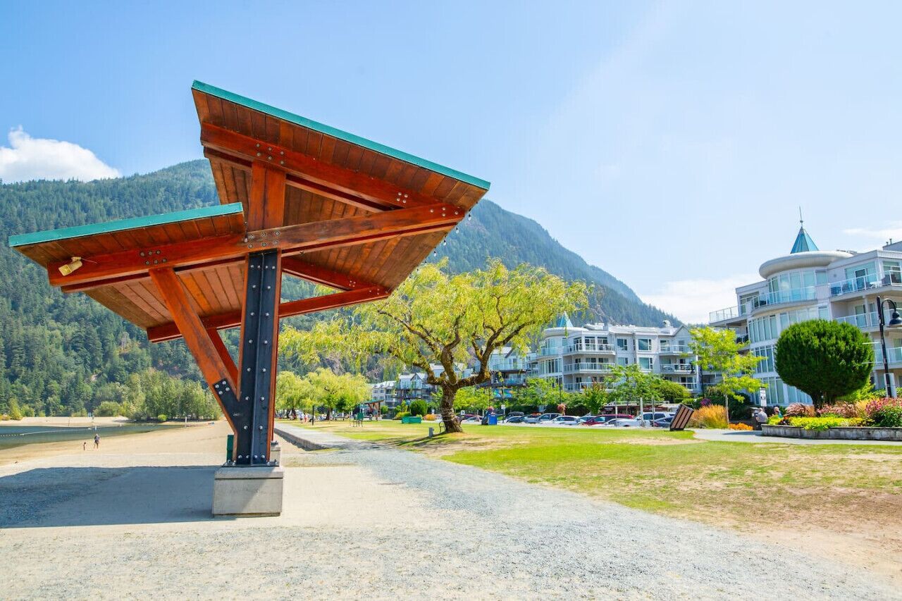A wooden pavilion with a green roof in a park with a mountain backdrop and modern buildings.