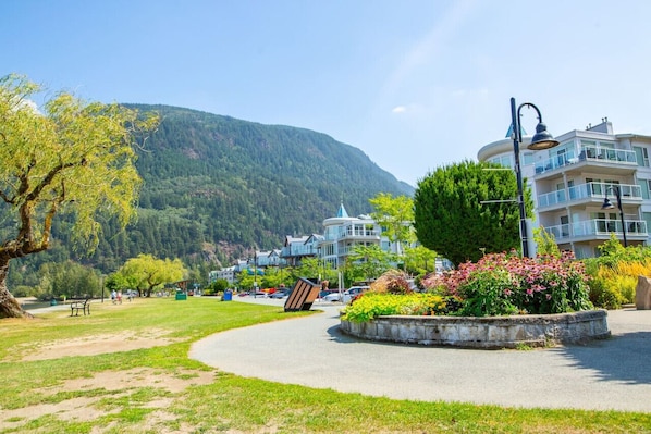 A park with a curved pathway, benches, and a flower bed. In the background, there is a building with a dome and balconies.