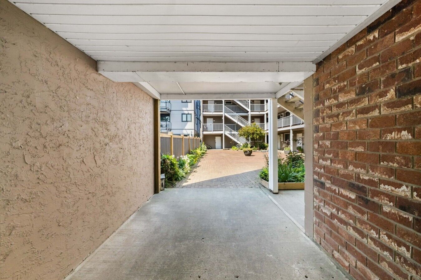 A corridor with a concrete floor, a brick wall on the right, and a building with balconies in the background.