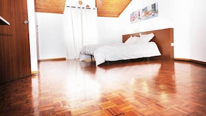 Un dormitorio con piso de madera, una cama con ropa de cama blanca, cabecera de madera y cortina blanca.