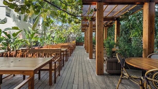 An outdoor wooden deck with tables and chairs, surrounded by lush greenery and a wooden pergola structure. - Global Luxury Suites at The Apex Miami (Miami Beach)