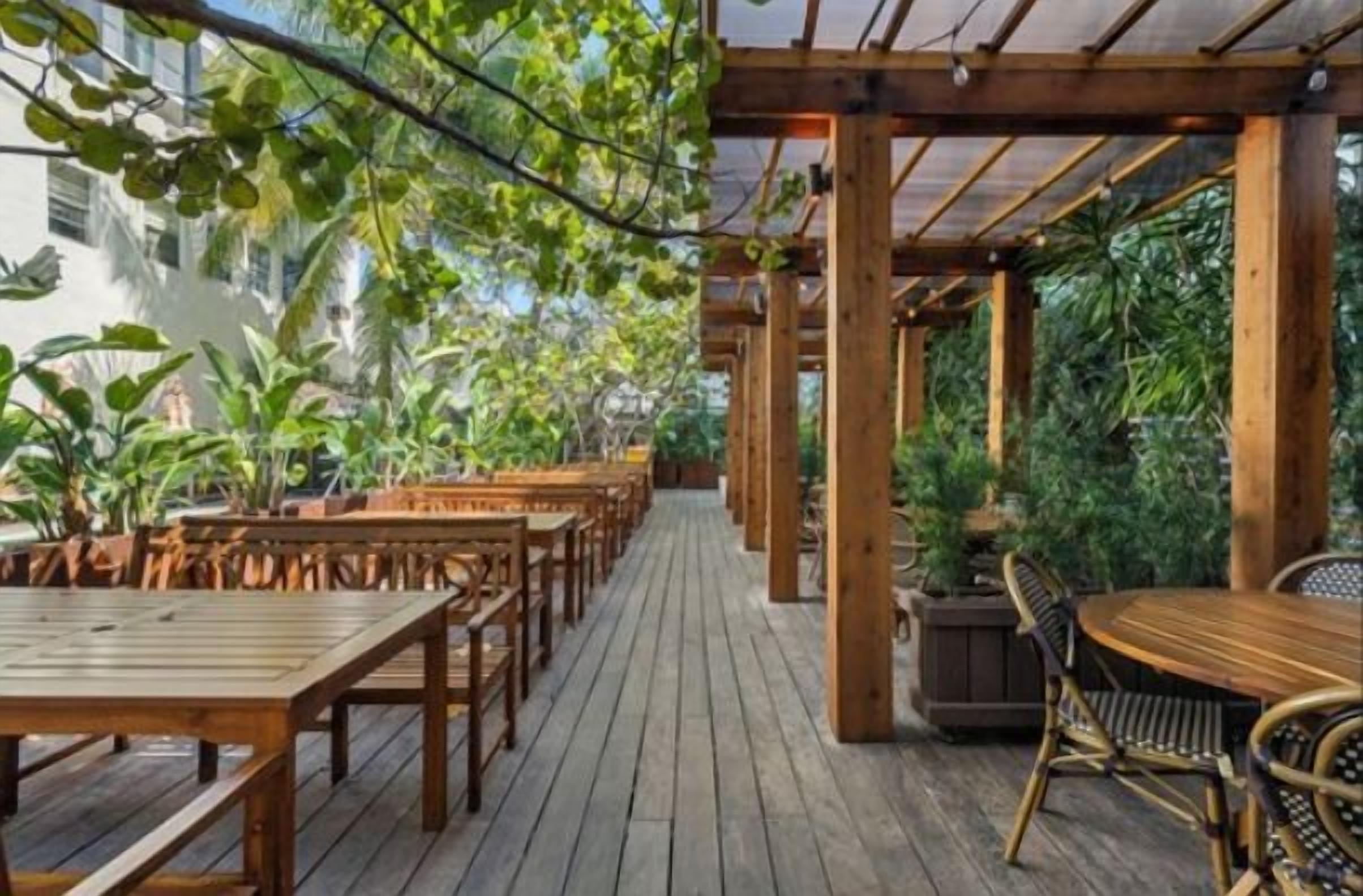 An outdoor wooden deck with tables and chairs, surrounded by lush greenery and a pergola structure.