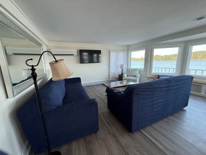 Un salon moderne avec un canapé, une table basse et une lampe. Un téléviseur est fixé au mur et une fenêtre offre une vue sur l’eau.