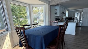 A dining area with a table set for six, a kitchen in the background, and large windows. - Heron's Landing (Harpswell)