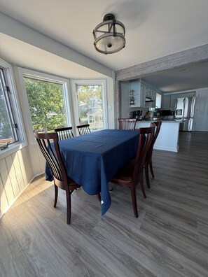 A dining area with a table set for six, a kitchen in the background, and large windows. - Heron's Landing (Harpswell)