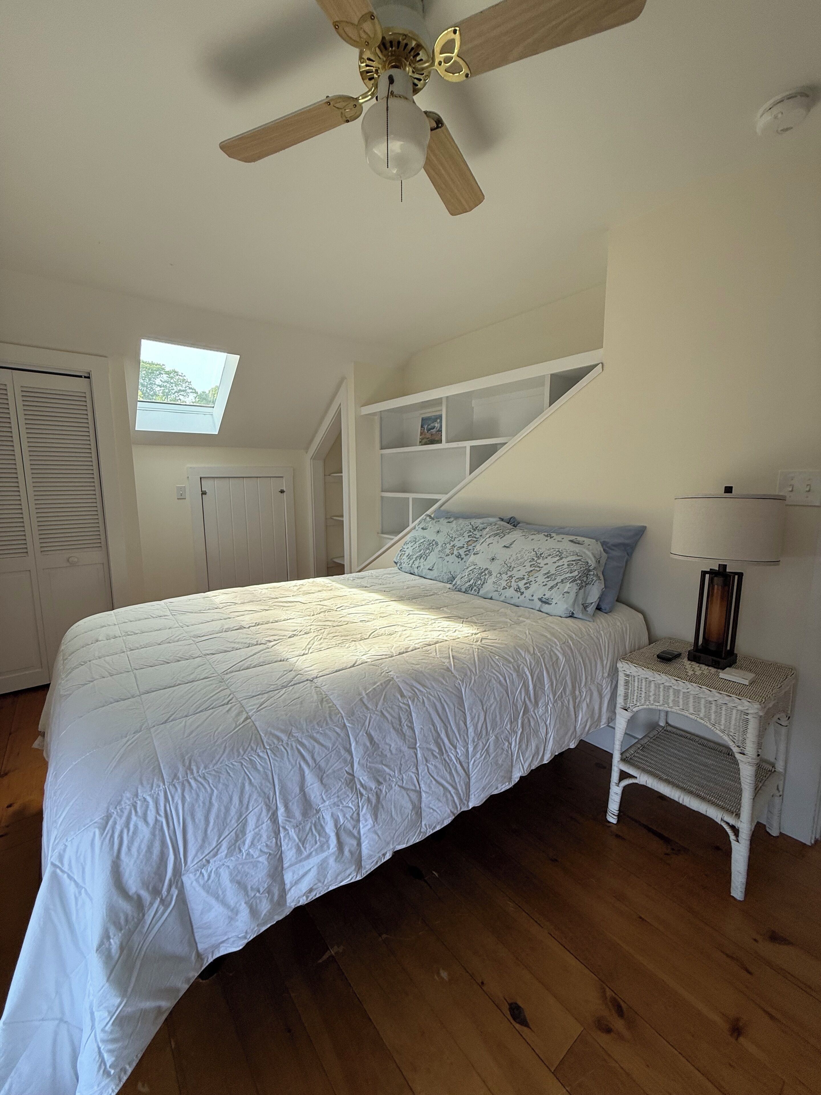 Una camera da letto con un letto, un comodino, una lampada, una ventola a soffitto, un lucernario e un'armadio bianco.