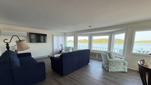 A living room with a sofa, armchair, and a table. There are windows with a view of the water and trees. - Heron's Landing (Harpswell)